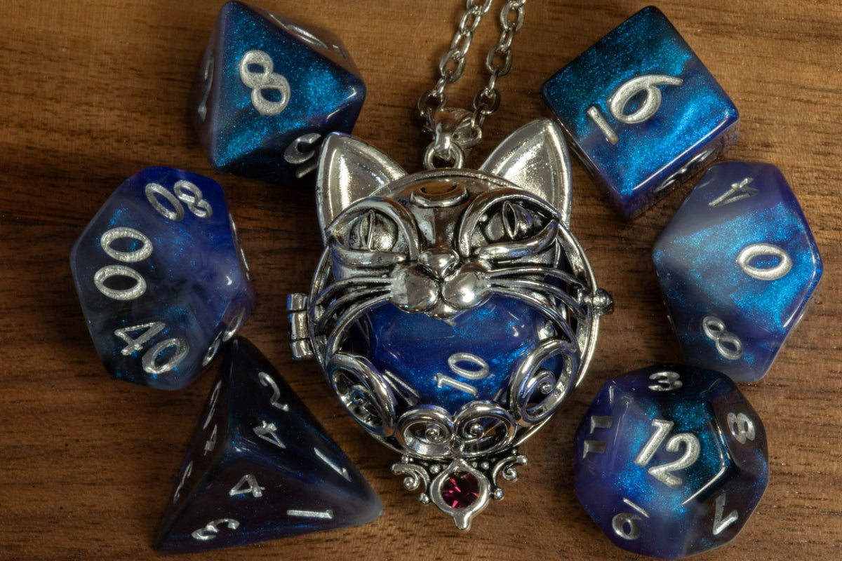 Cat pendant in a silver finish with a pink crystal, holding a blue galaxy D20, beside a blue galaxy dice set on wood