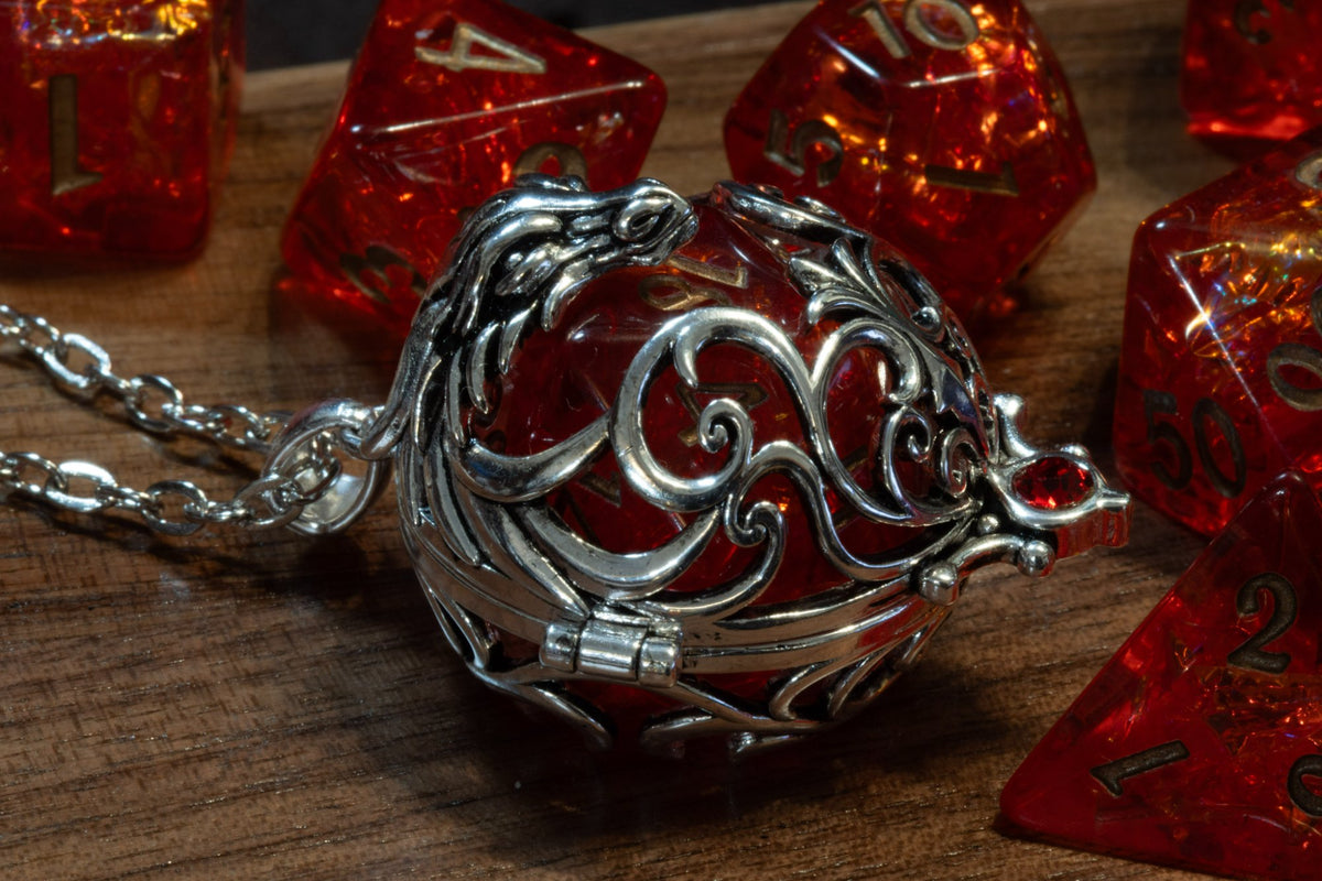Dragon pendant in a silver finish, closed, with a red D20 inside, lying on wood beside red holographic dice