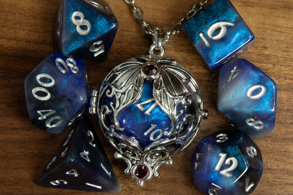 Lunar moth pendant in a silver finish with two purple crystals, holding a blue galaxy D20, beside a blue galaxy dice set on wood