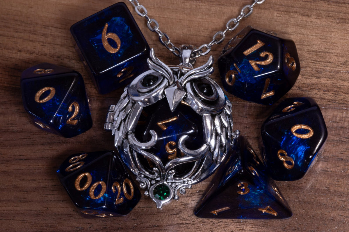 Owl pendant in a silver finish with a green crystal, holding a dark blue D20, beside a dark blue dice set with gold numbers on wood