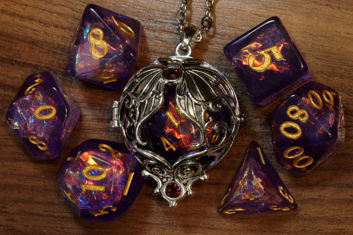 Lunar moth pendant in a silver finish with two red crystals, holding a purple holographic D20, beside a purple holographic dice set with gold numbers on wood