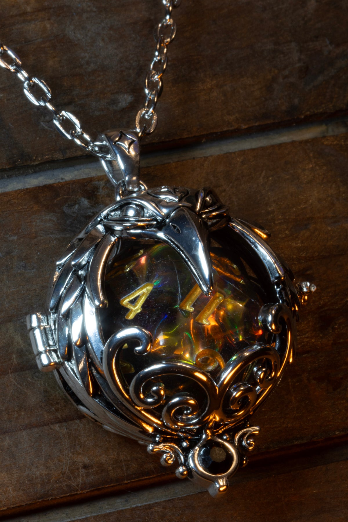 Raven pendant in a silver finish with a black crystal, holding a clear holographic D20 with gold numbers, hanging on its chain against wood